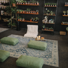 Load image into Gallery viewer, Room with shelves filled with plants, green cushions on a rug, and candles.
